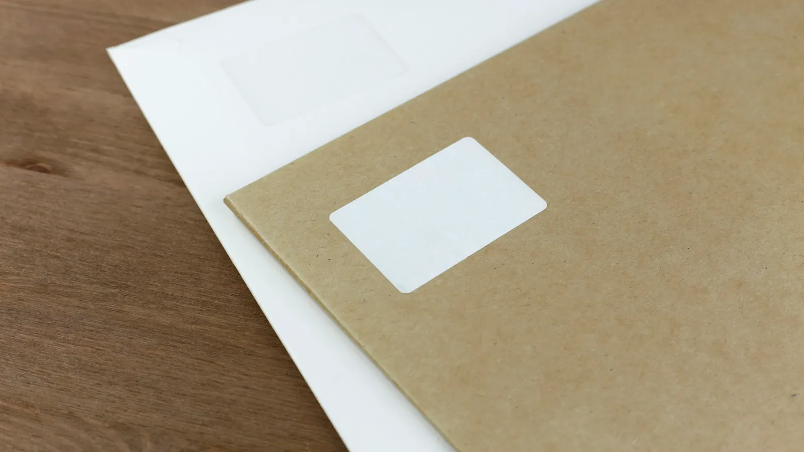 A kraft envelope with a blank address label on top of a white window envelope on a wooden table
