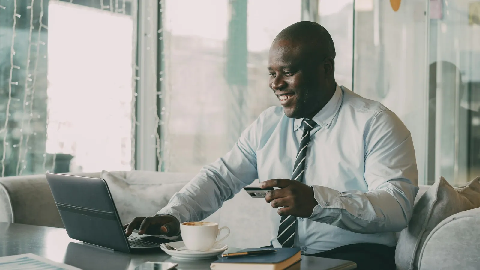 Man in a shirt and tie holding a payment card while typing on a laptop at a café table