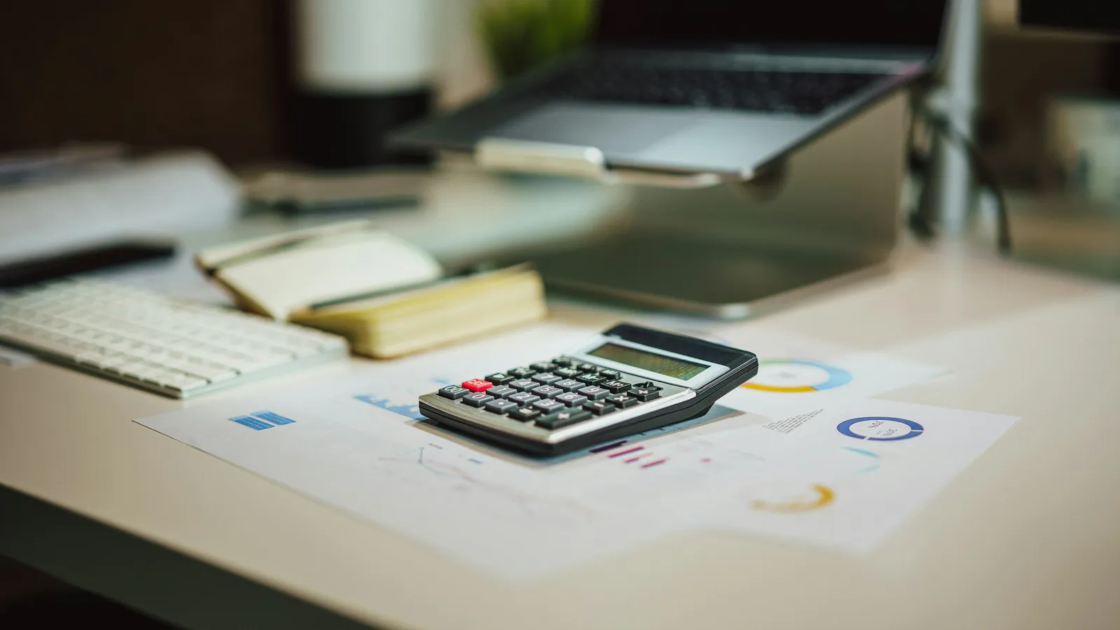 Calculator on printed charts on a desk, with a laptop on a stand behind it