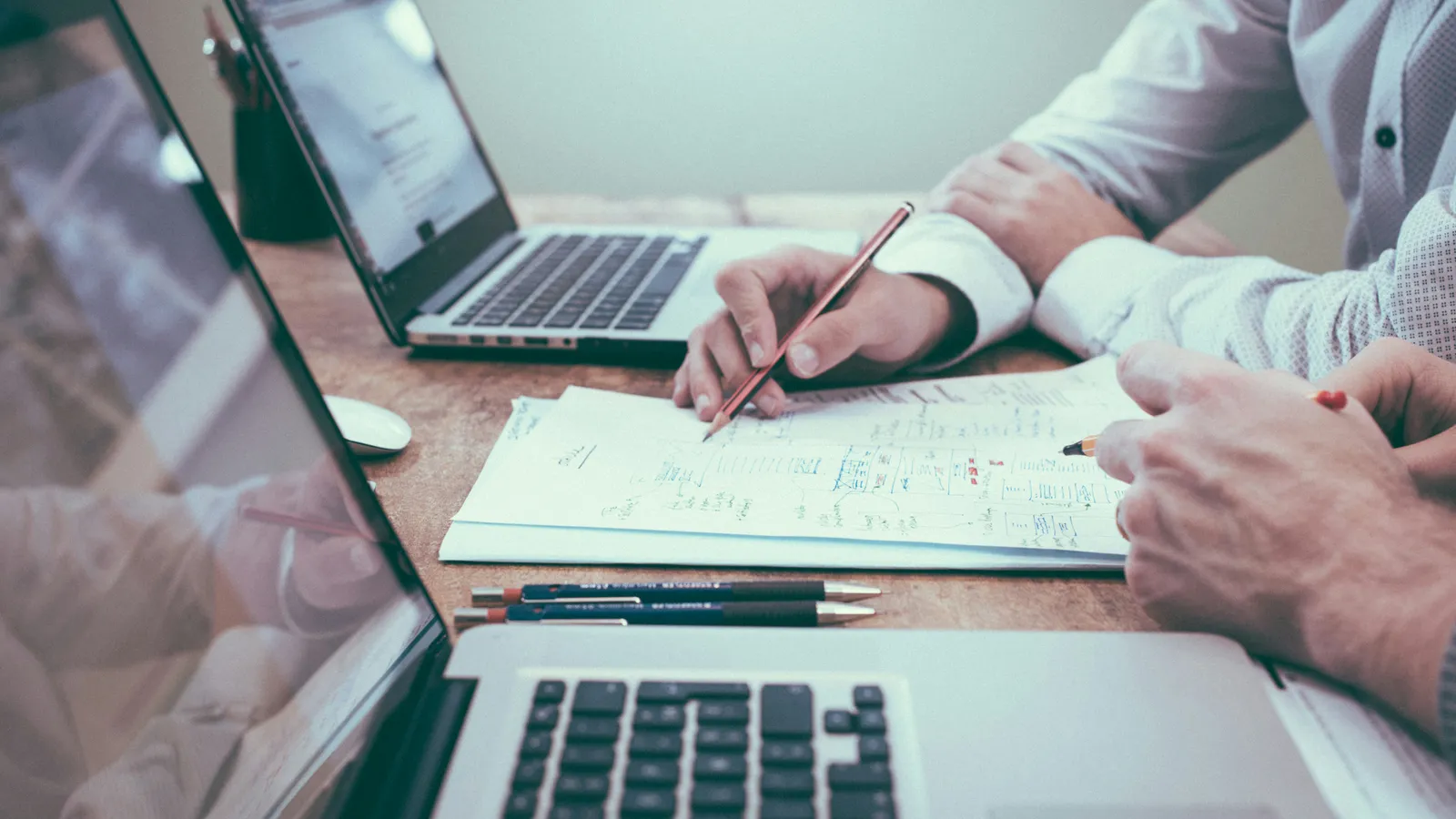 Two people reviewing handwritten business paperwork with a pencil between two open laptops