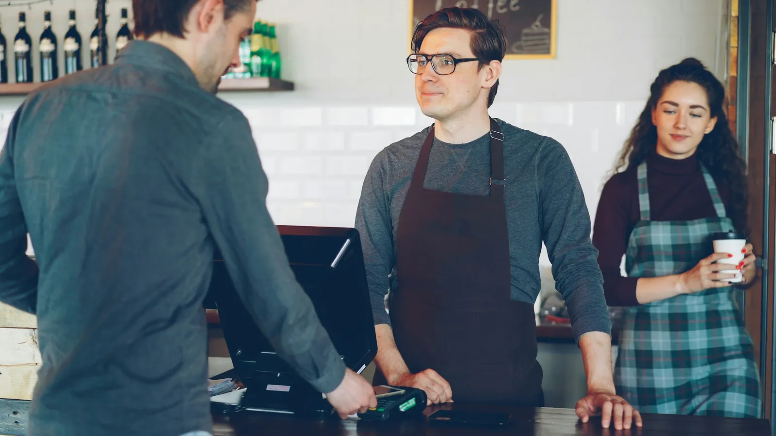Customer paying at a cafe counter terminal while two staff in aprons look on