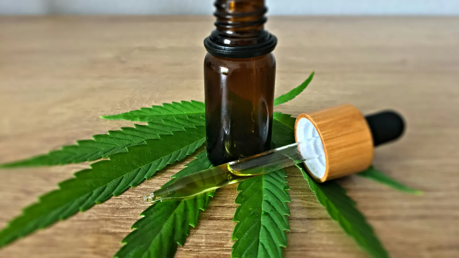 Amber glass dropper bottle and its dropper resting on a green hemp leaf on a wooden table