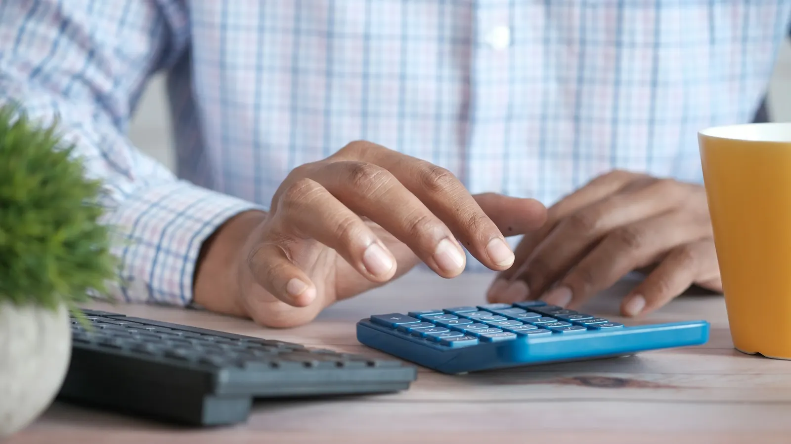 Hands using a blue calculator on a desk next to a keyboard, a yellow mug and a small plant