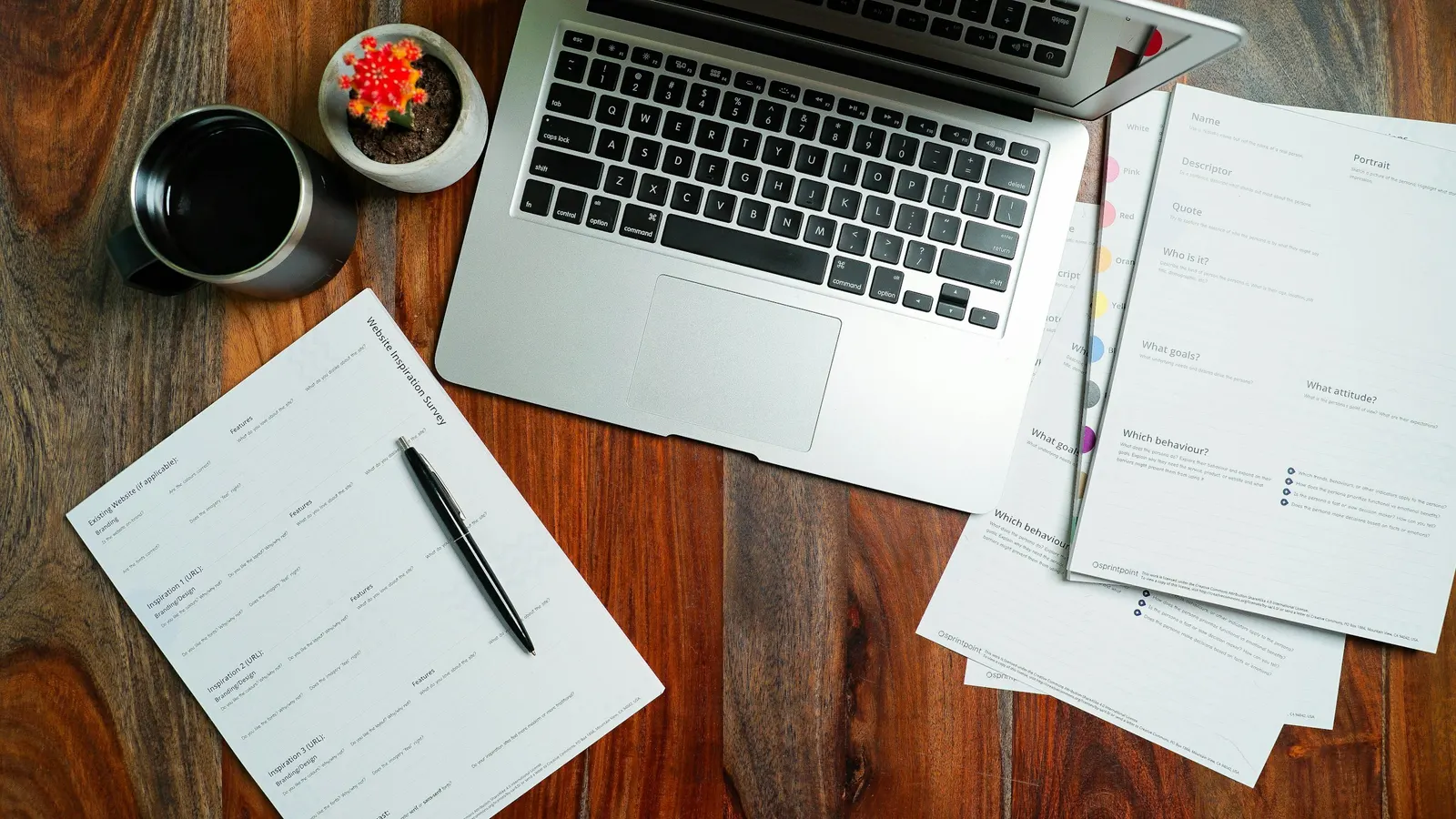 Overhead view of an open laptop, printed forms with a pen, a coffee mug and a small cactus on a wooden desk