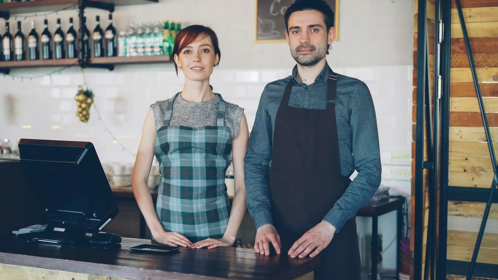 Two café workers in aprons standing behind a counter next to a register screen
