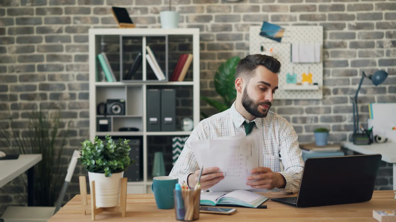 Man reading printed papers at a desk with an open laptop
