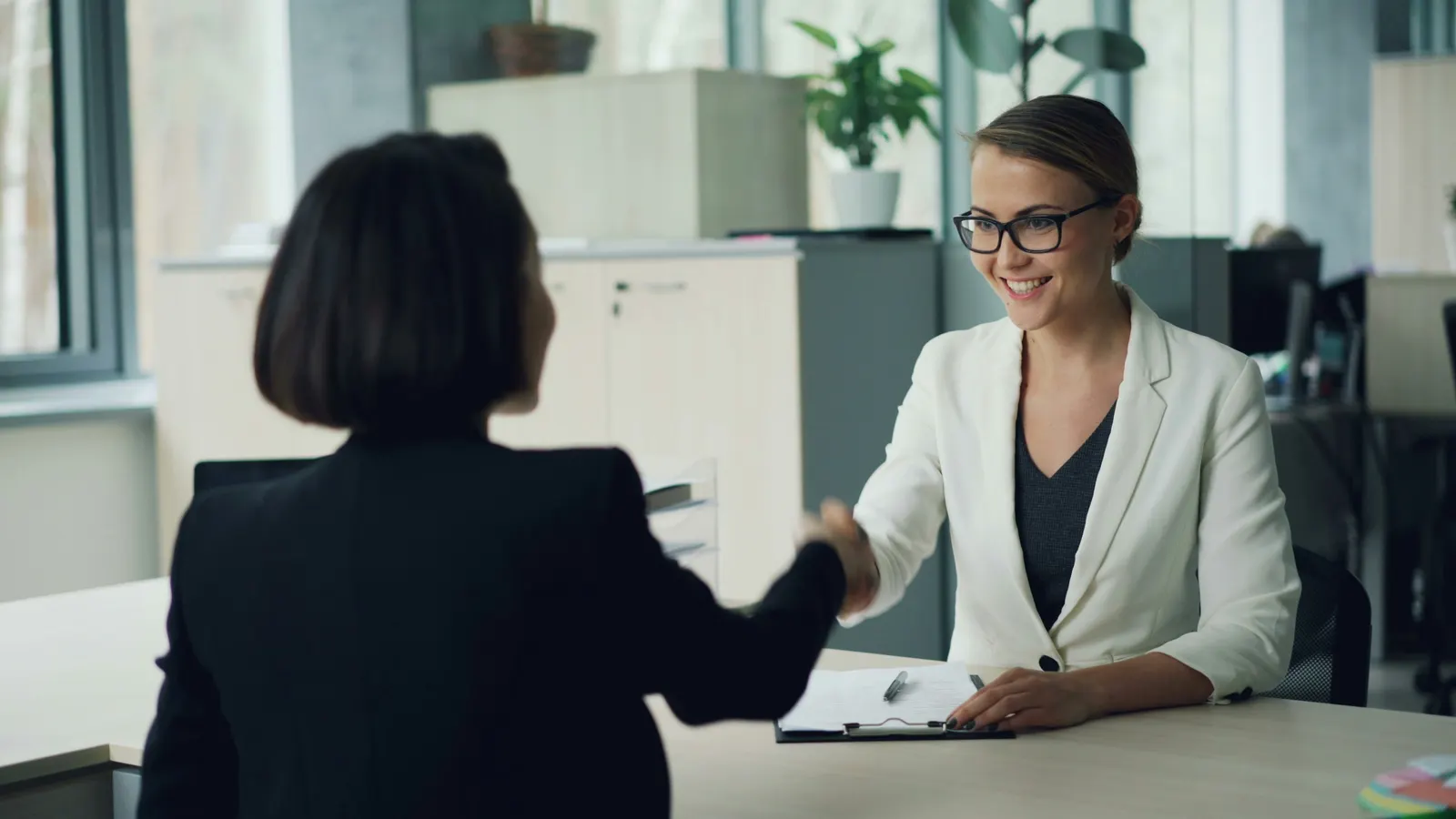 Two women shaking hands across a desk in a bright office