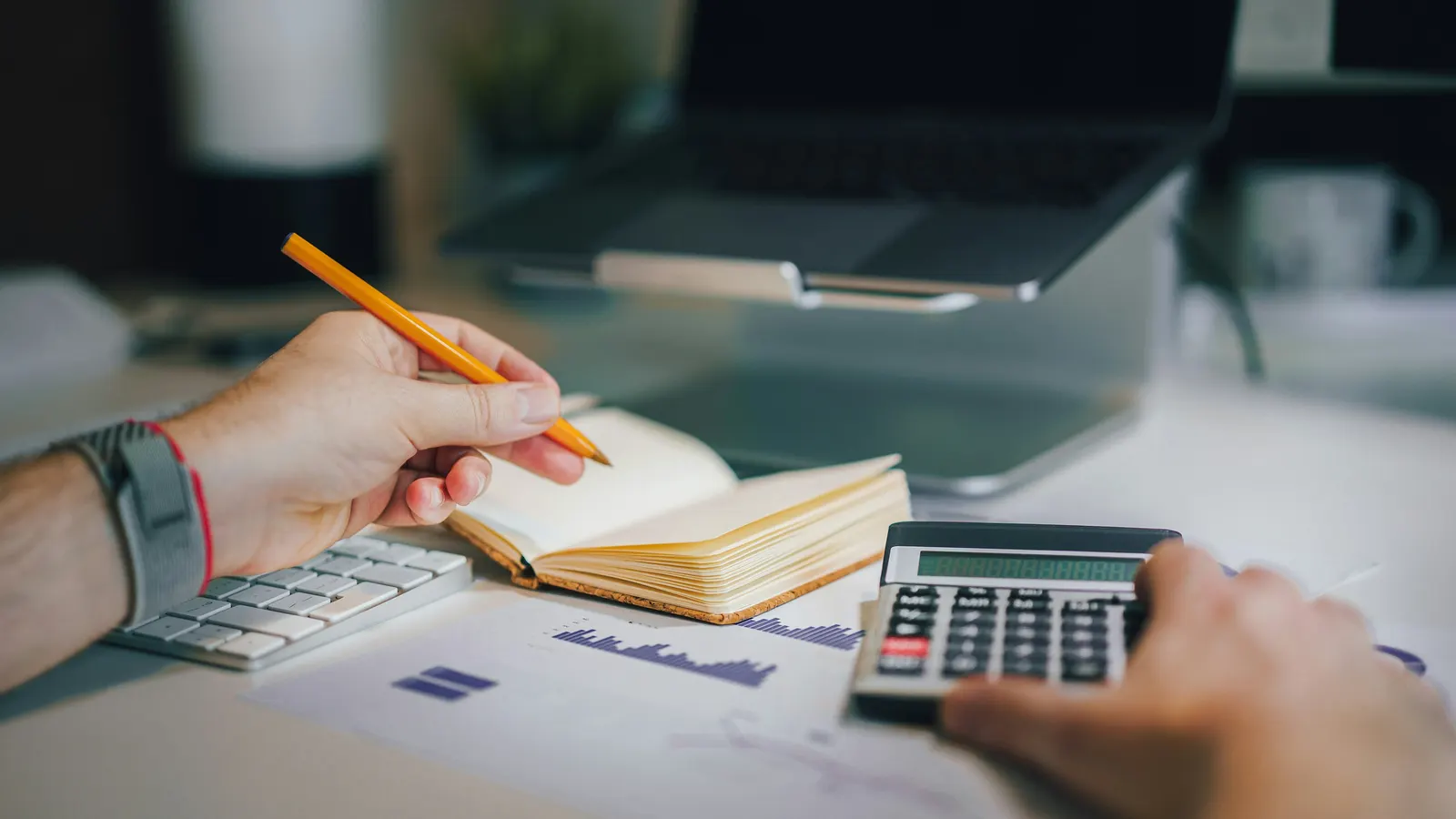 Hands holding a pencil and a calculator over a notebook and printed charts on a desk