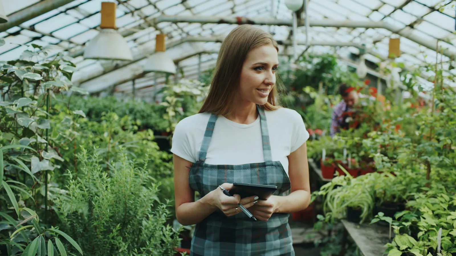 Woman in a plaid apron holding a tablet and a pen in a greenhouse full of plants