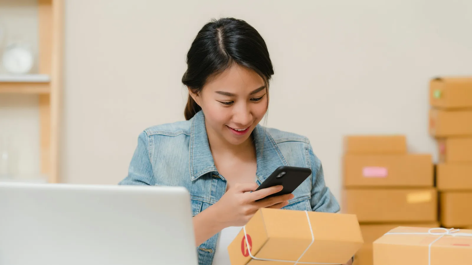 Woman checking a parcel with her phone beside a laptop and stacked shipping boxes