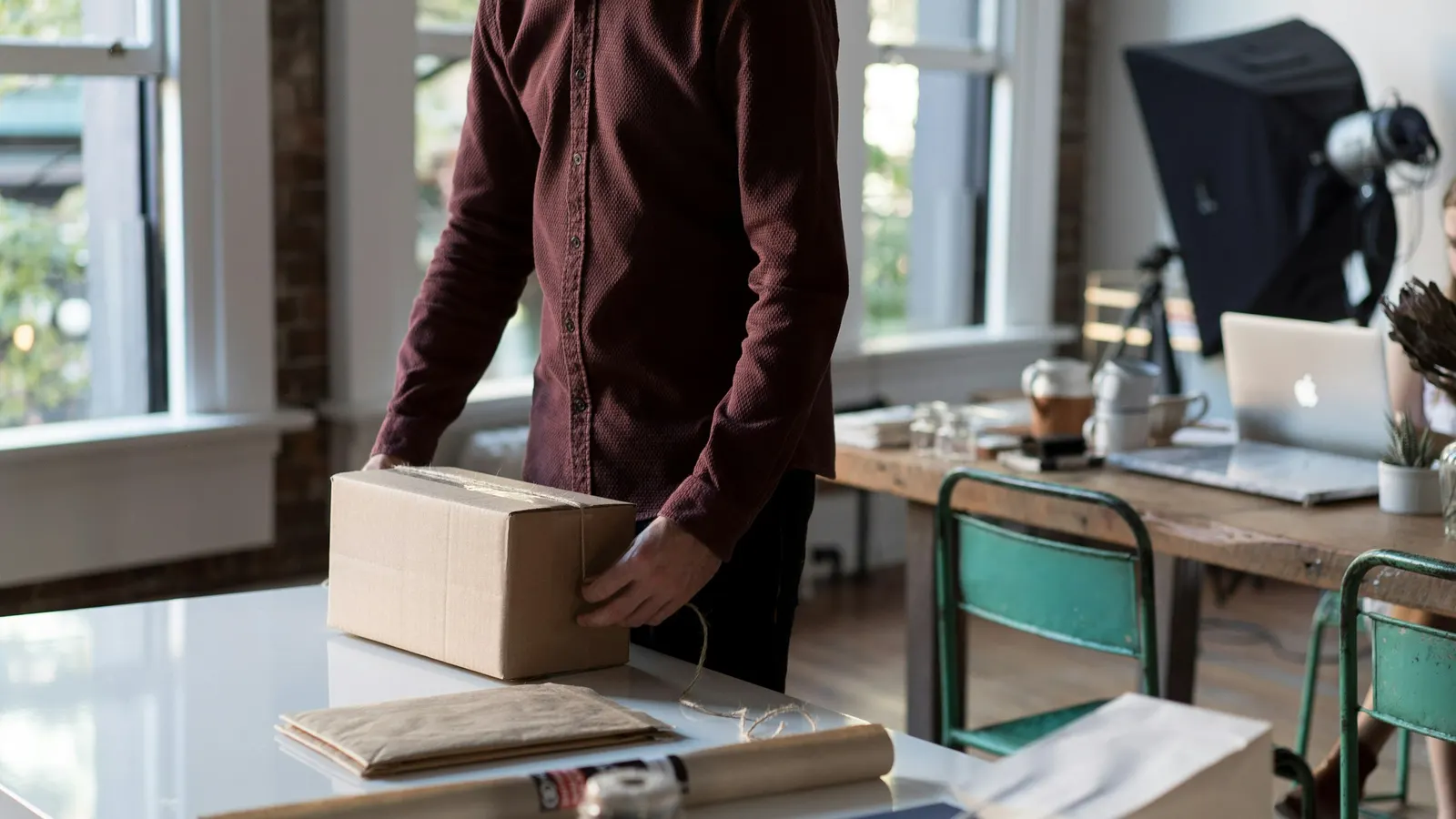 Person packing a cardboard shipping box on a white table, with a laptop on a desk behind them