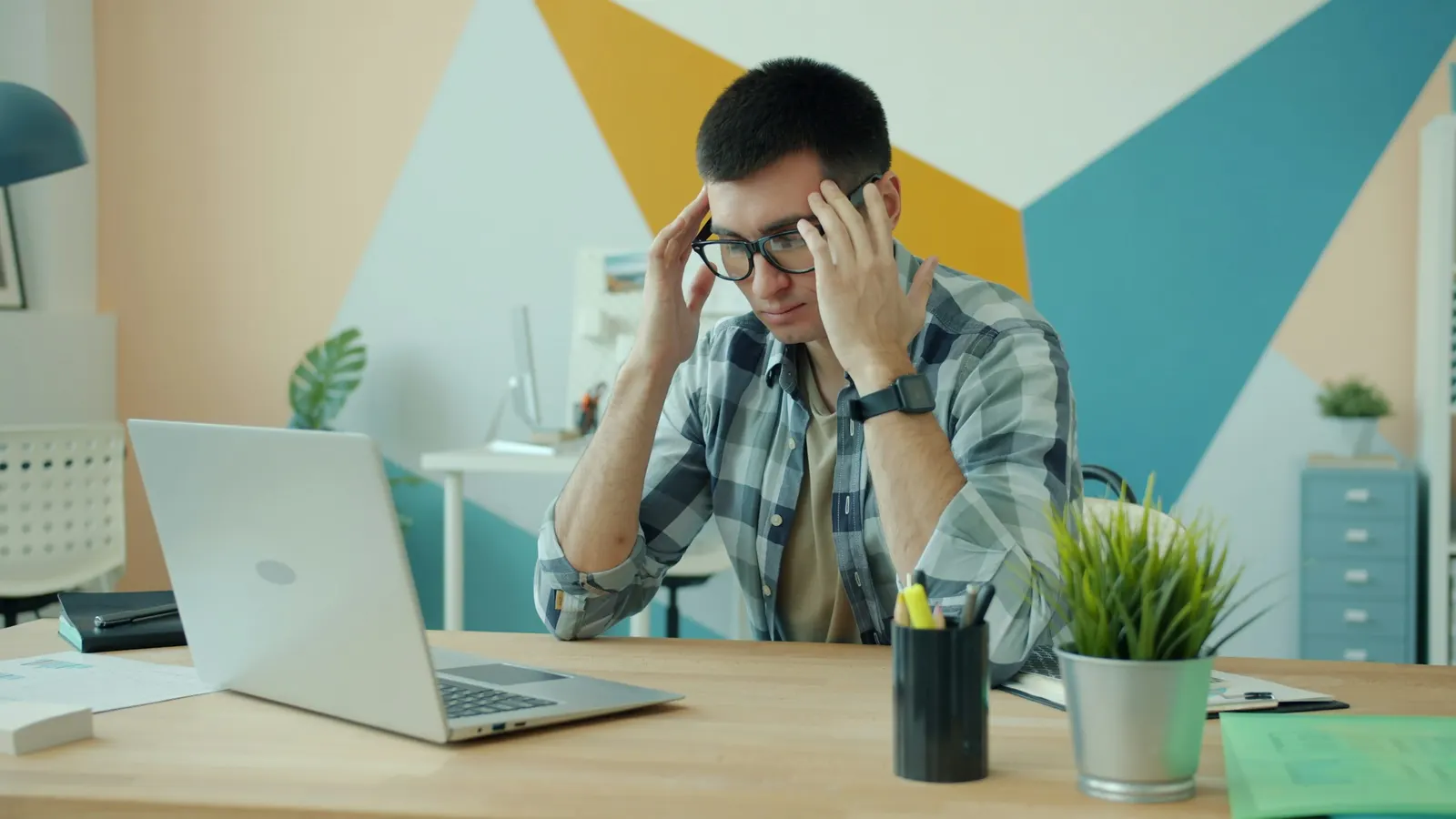 A man at a desk with a laptop, holding his head in both hands